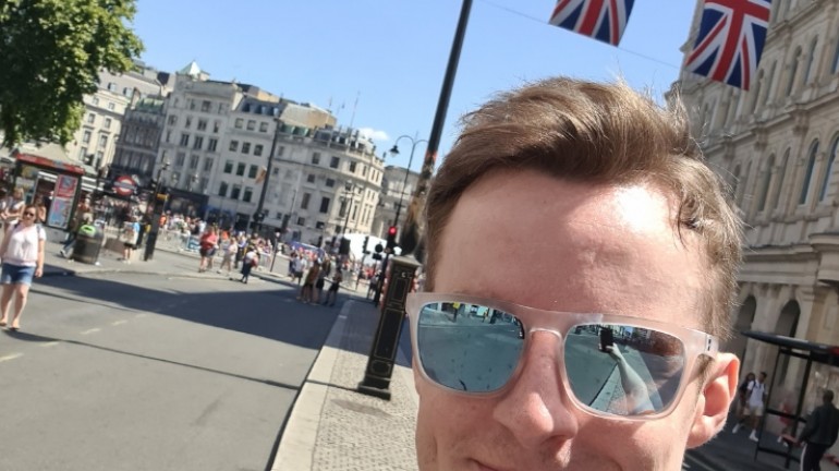 A picture of myself (Luc Shelton) posing with my medal after running the ASICS London 10K. Behind me is a string of Union Jack flags hanging across the street next to Trafalgar Square.